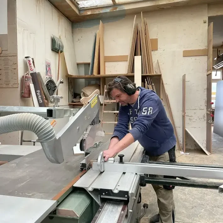 Ein Schreiner arbeitet mit einer Tischsäge in einer Werkstatt. Holzstücke liegen bereit.